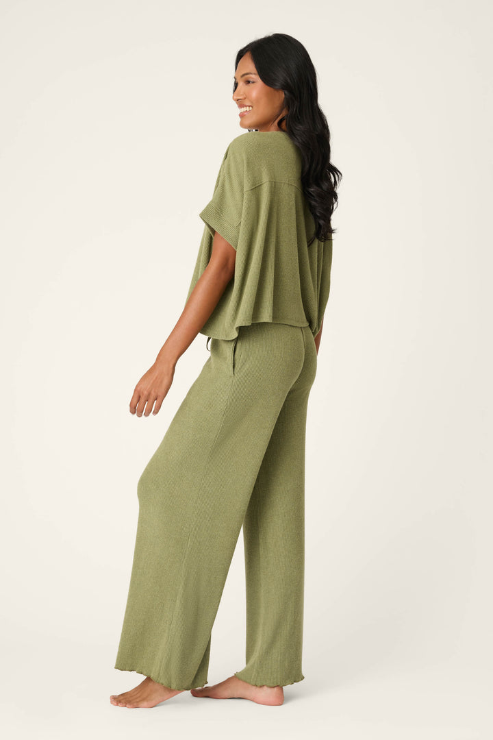 A woman with long dark hair smiles barefoot in the P.J. Salvage Olive You Forever Lounge Set, featuring a loose short-sleeve olive top and matching wide-leg pants, shown from side and back against a plain light background.