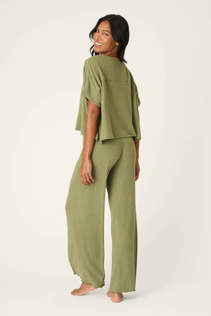 A woman smiles barefoot, glancing over her shoulder in the P.J. Salvage Olive You Forever Lounge Set—an olive green, loose short-sleeve top with matching wide-leg pants—against a plain light background.