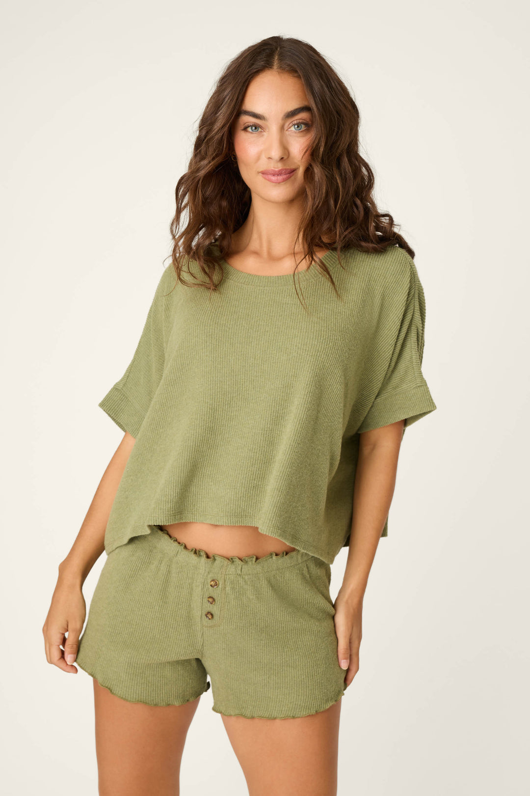A woman with long brown hair wears the P.J. Salvage Olive You Forever Lounge Short Sleeve Top and matching buttoned shorts, standing against a light background and smiling softly at the camera.