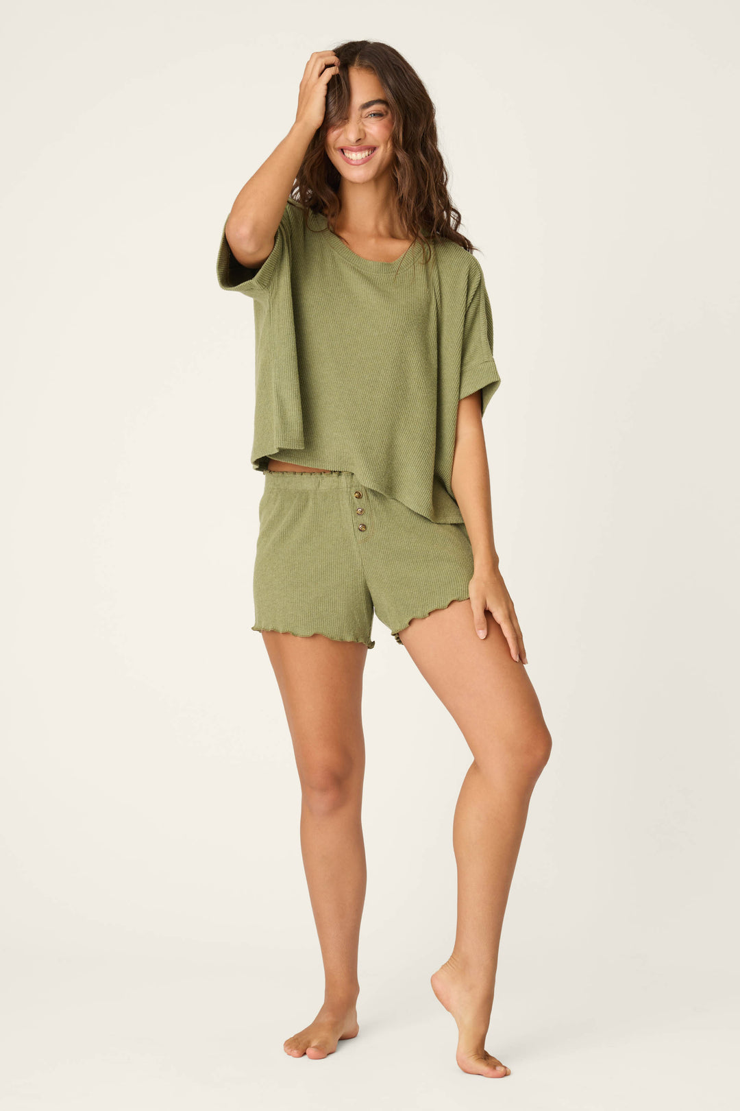 A woman with long brown hair smiles and poses barefoot, wearing the P.J. Salvage Olive You Forever Lounge Short Sleeve Top and matching shorts against a plain light background.