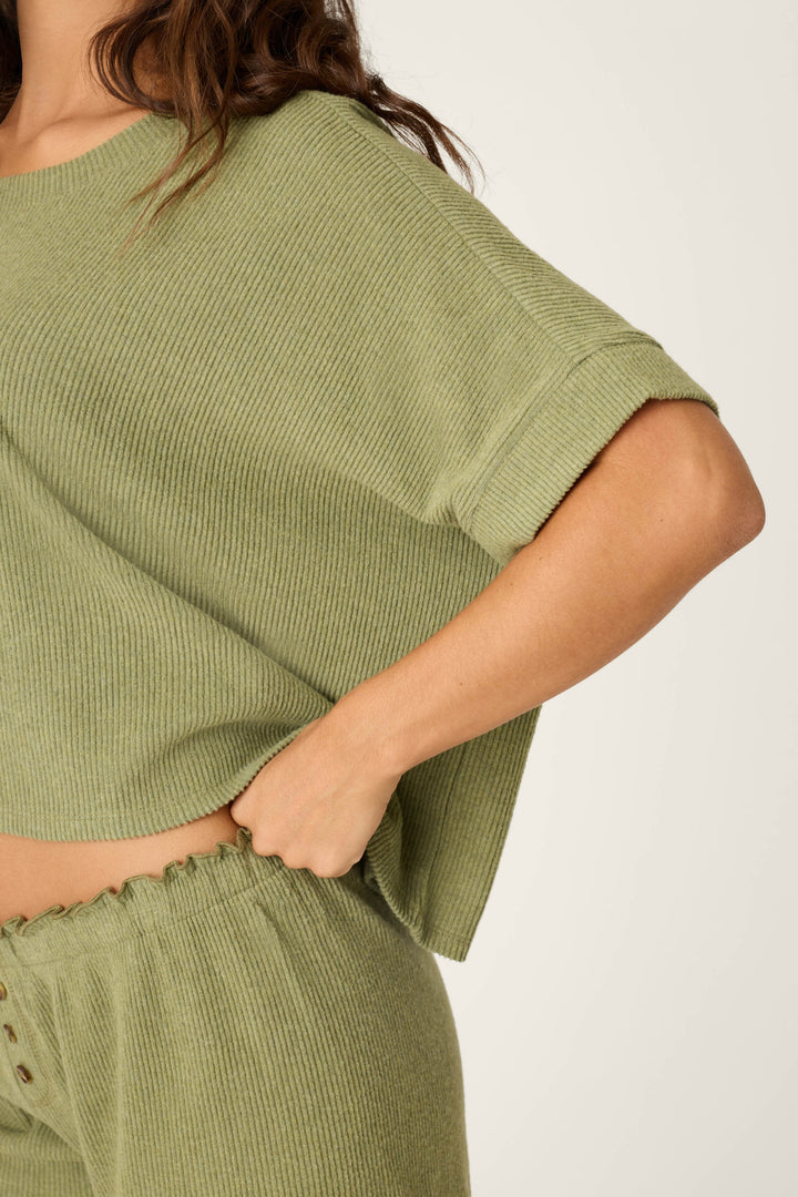 A person models the P.J. Salvage Olive You Forever Lounge Short Sleeve Top, a loose ribbed light green shirt paired with matching high-waisted pants, shown from shoulders to mid-thigh with one hand on their hip.