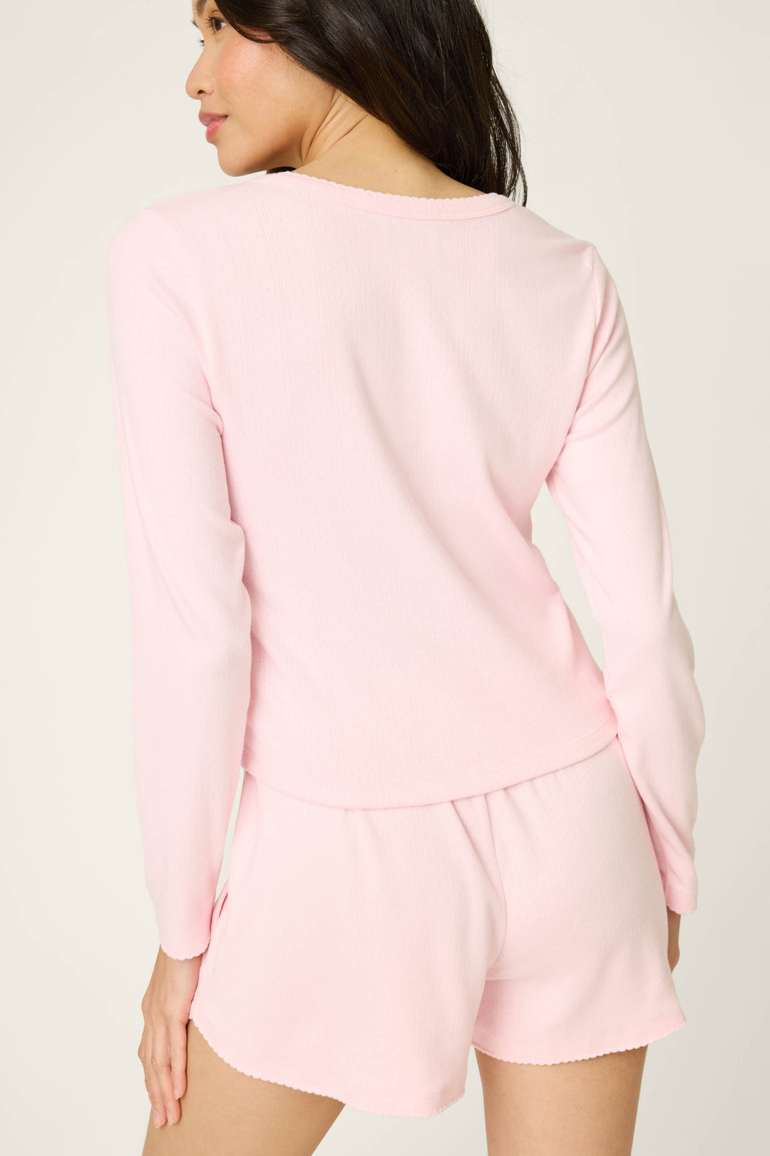 A woman stands with her back to the camera wearing the P.J. Salvage Pointelle Essentials Ballet Pink Cardigan, styled with matching shorts. The soft, comfortable outfit is set against a plain, light-colored background.