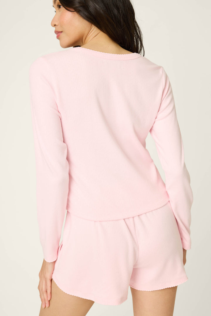 A woman stands with her back to the camera wearing the P.J. Salvage Pointelle Essentials Ballet Pink Cardigan, styled with matching shorts. The soft, comfortable outfit is set against a plain, light-colored background.