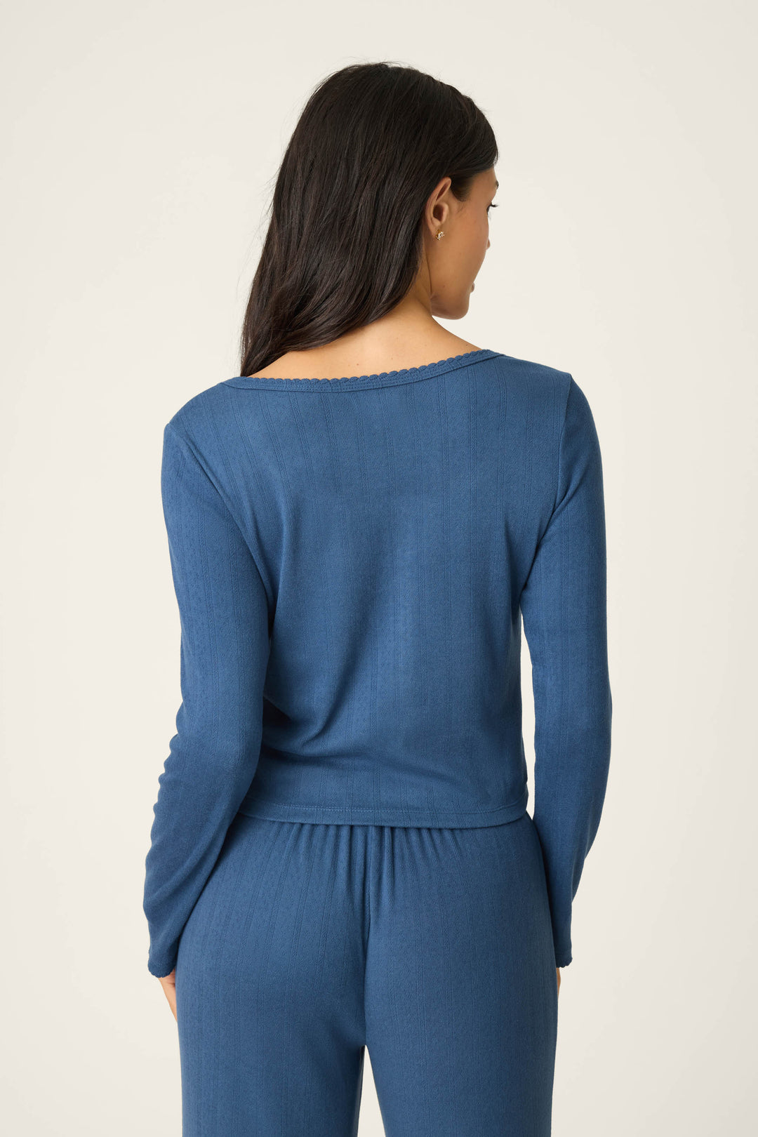 A woman with long dark hair faces away from the camera, wearing the P.J. Salvage Pointelle Essentials Deep Sea Cardigan lounge set. She stands against a plain, light-colored backdrop.