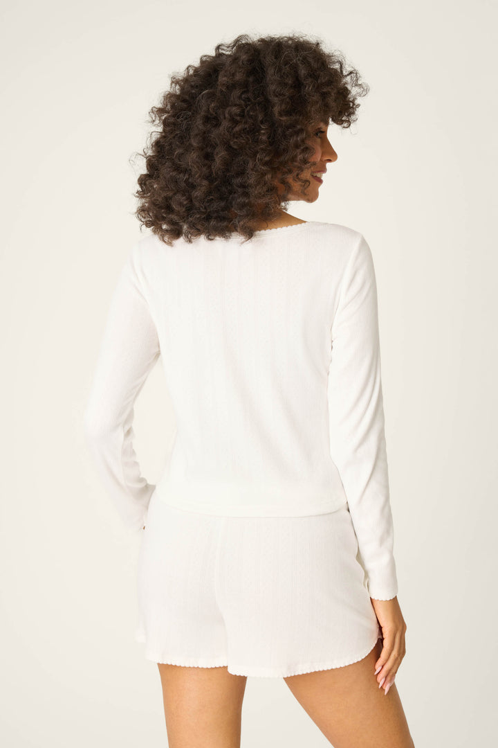 A person with curly hair stands with their back to the camera, wearing the P.J. Salvage Pointelle Essentials Ivory Cardigan. They pair it with matching white shorts against a plain, light-colored background.