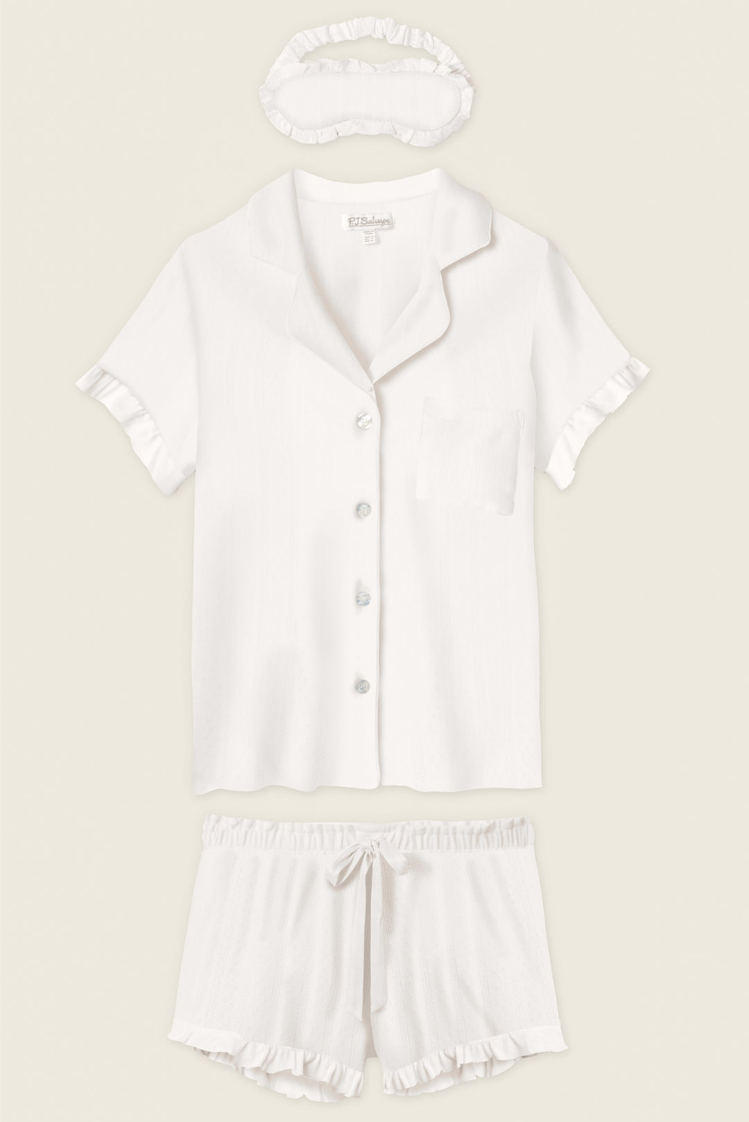 The P.J. Salvage Pointelle Essentials Ivory 3-Piece Shorts Set includes a short-sleeve button-up shirt with chest pocket, matching drawstring shorts, and a sleep mask—all in white with ruffle trim.