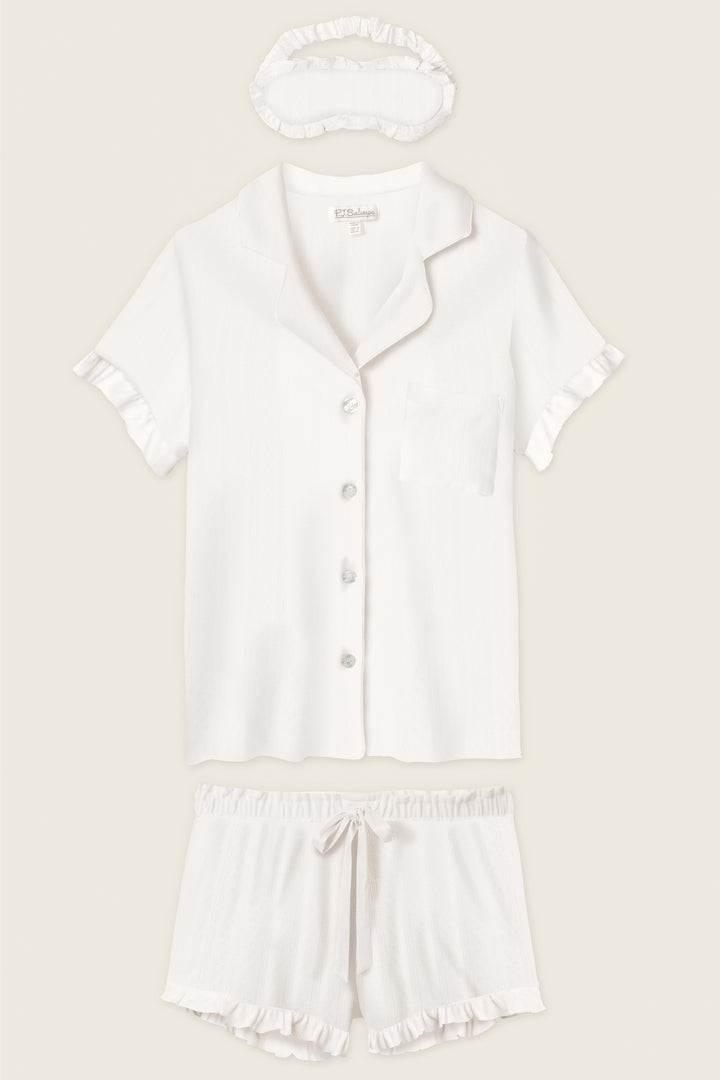 The P.J. Salvage Pointelle Essentials Ivory 3-Piece Shorts Set includes a short-sleeve button-up shirt with chest pocket, matching drawstring shorts, and a sleep mask—all in white with ruffle trim.