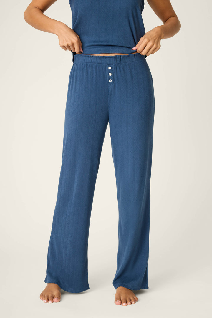 A person stands barefoot adjusting the waistband of P.J. Salvage Pointelle Essentials Deep Sea Open Leg Pants—loose-fitting, high-waisted lounge pants in blue with an adjustable drawstring and three front buttons, paired with a matching sleeveless top.