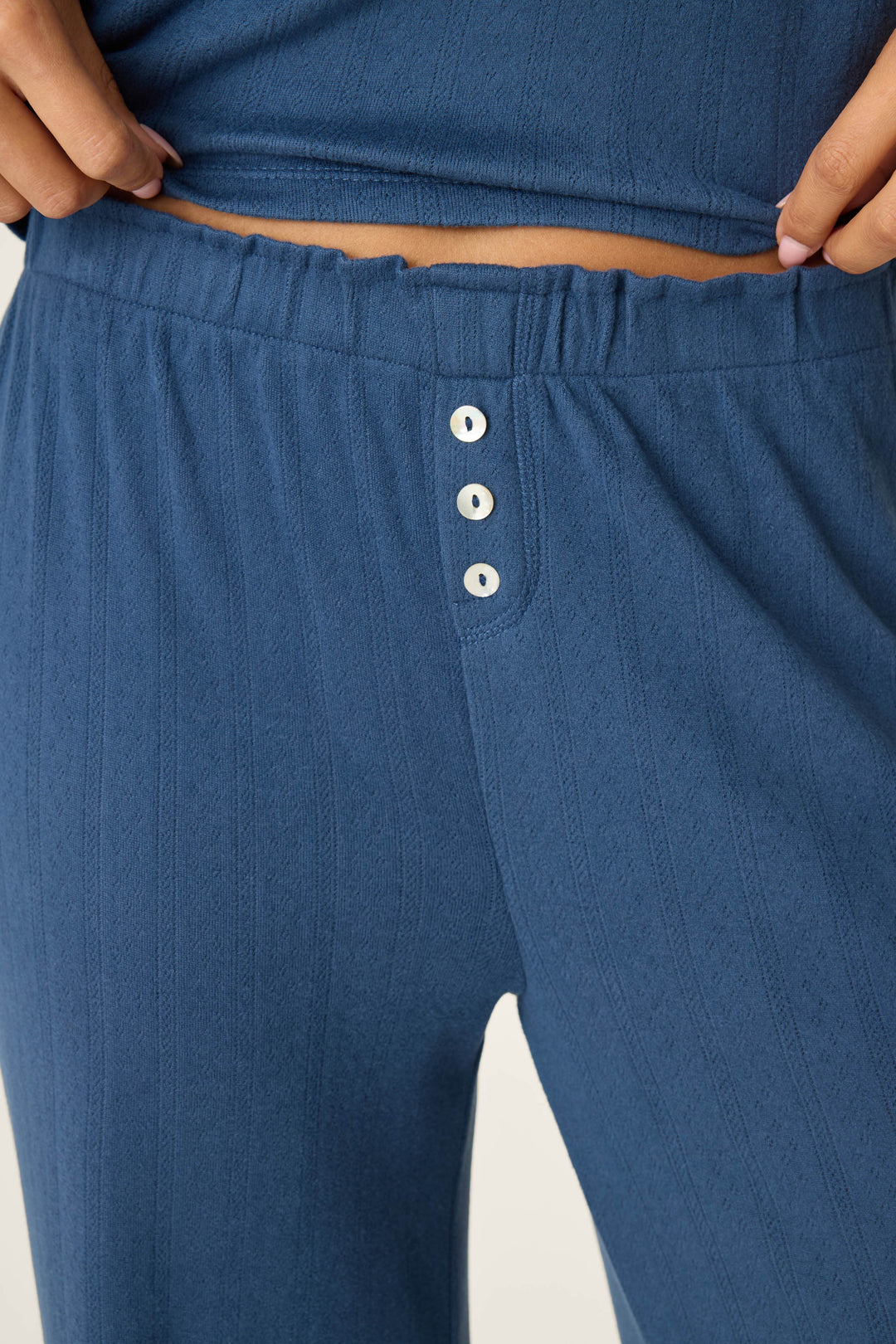 A person is shown adjusting the adjustable drawstring on their P.J. Salvage Pointelle Essentials Deep Sea Open Leg Pants, a blue ribbed lounge style featuring three white buttons on the front.