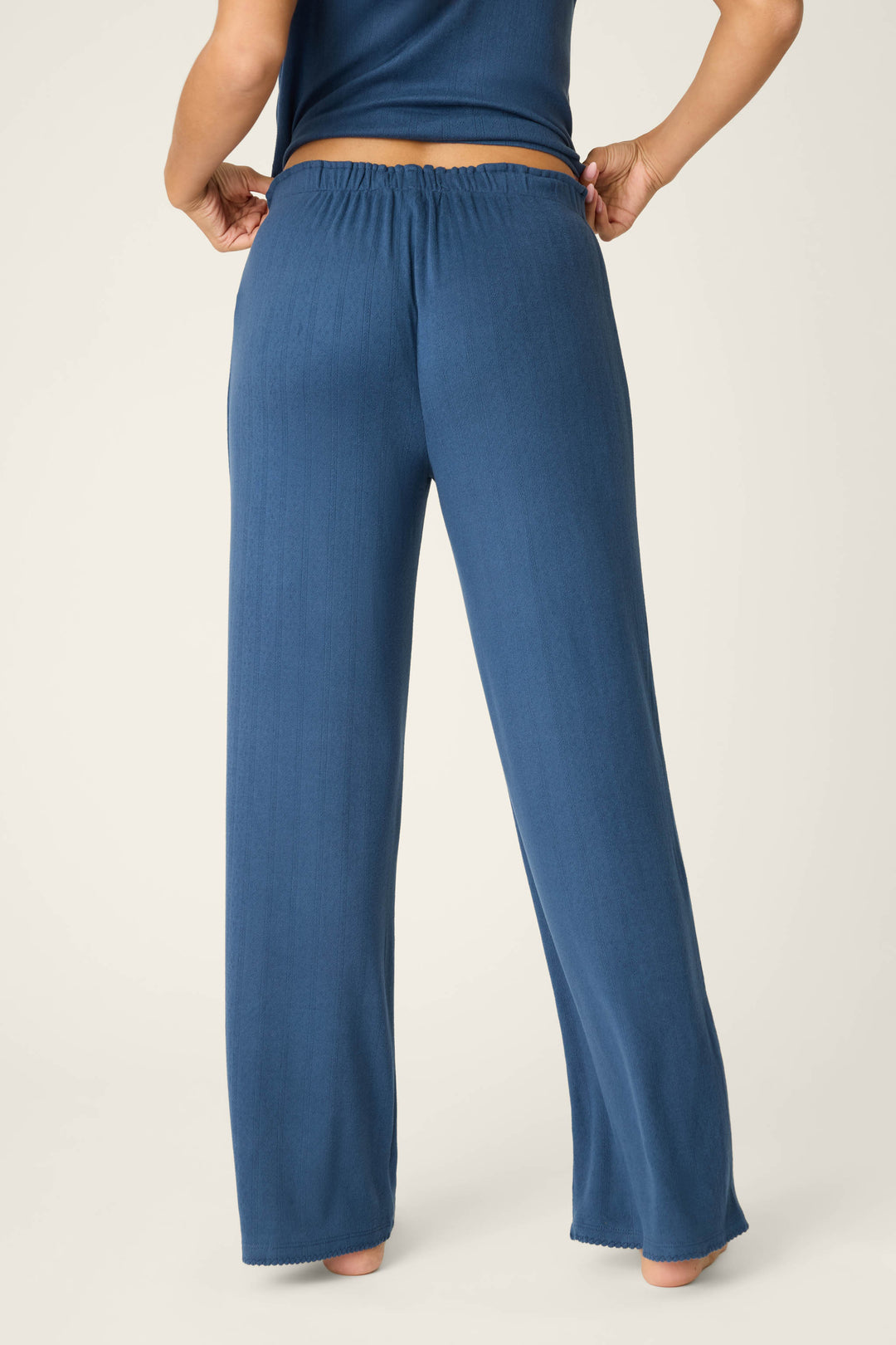 A barefoot person, shown waist down and facing away, wears P.J. Salvage Pointelle Essentials Deep Sea Open Leg Pants—loose-fitting, high-waisted blue lounge pants with an adjustable drawstring and a matching top.