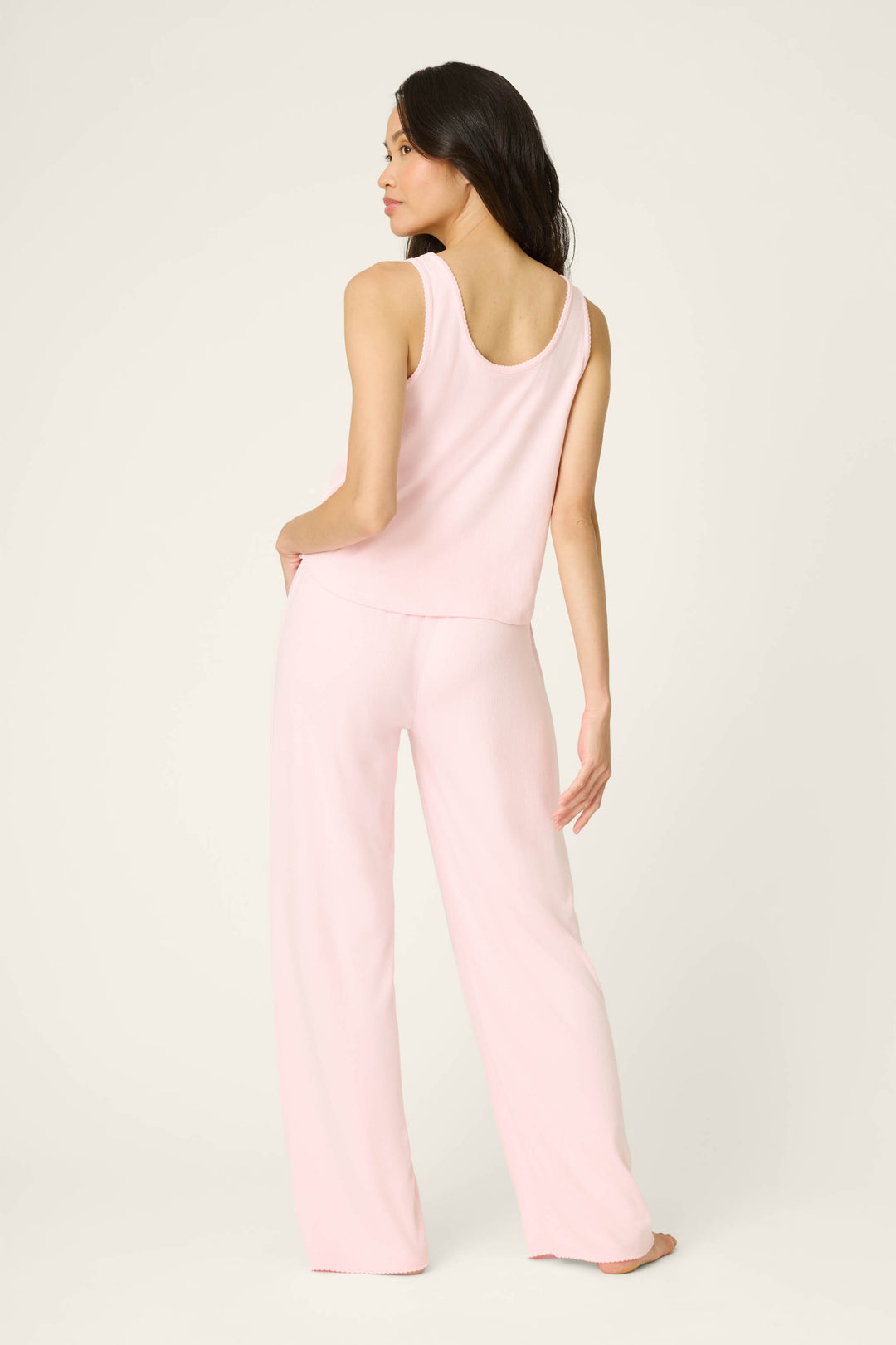 A woman with long dark hair stands barefoot, facing away against a light background, wearing the P.J. Salvage Pointelle Essentials Ballet Pink PJ Set—a sleeveless top and loose matching pants. One hand is on her hip, the other relaxed by her side.