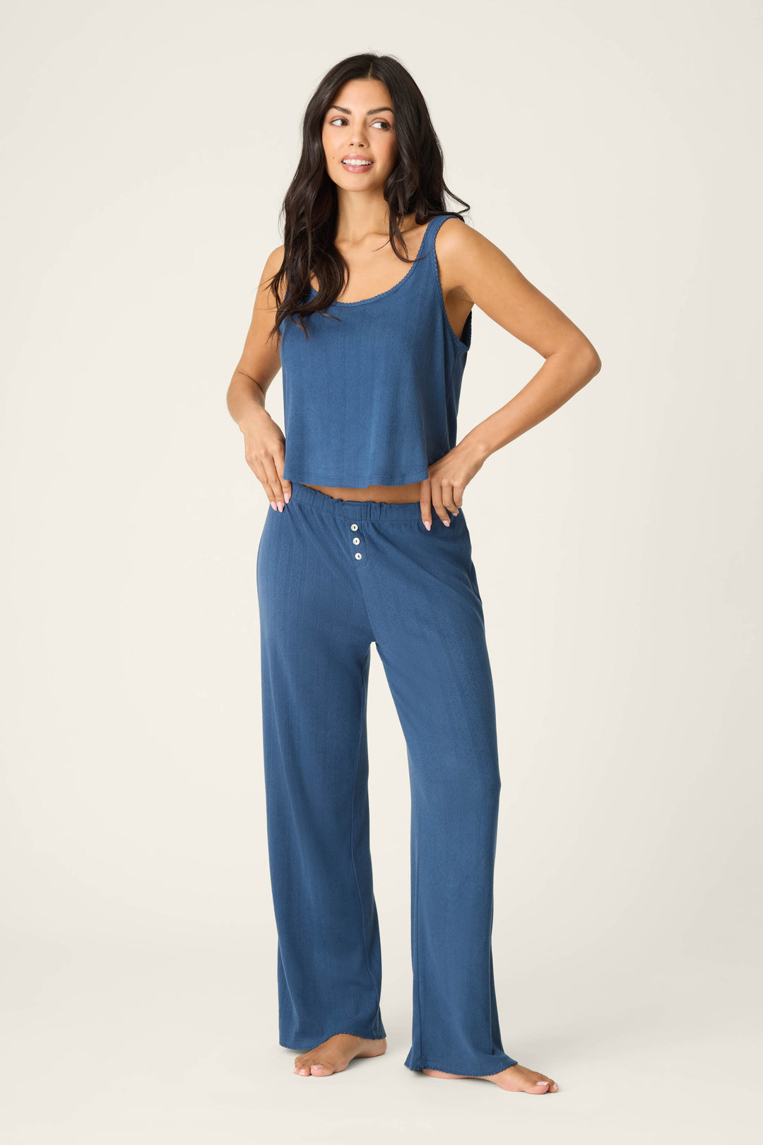 A woman stands barefoot against a plain backdrop, wearing the P.J. Salvage Pointelle Essentials Deep Sea PJ Set—a relaxed two-piece with a blue cropped tank pajama top and loose buttoned blue pants. She gazes to the side, hands resting on her waistband.