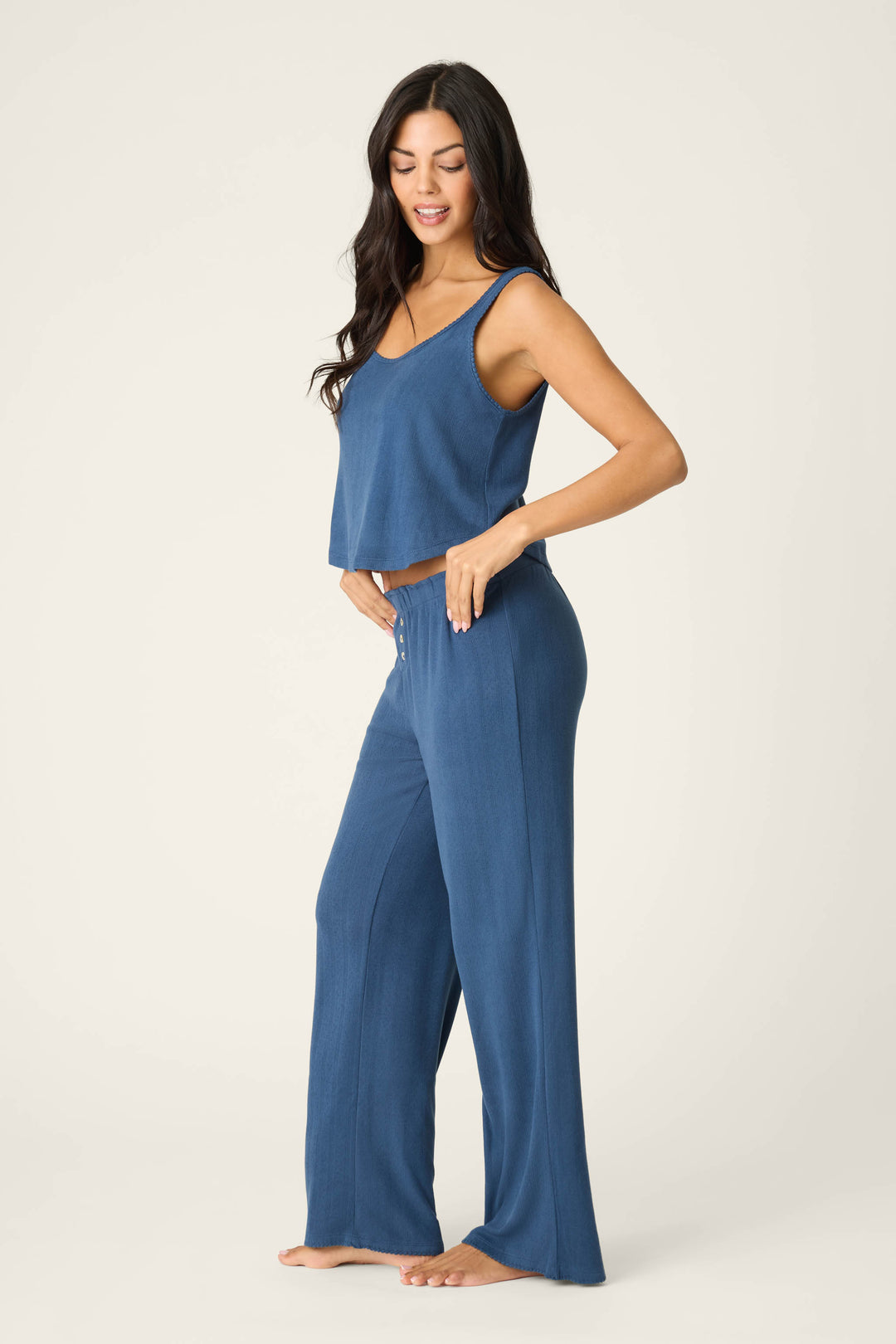 A woman stands barefoot against a plain background, wearing the P.J. Salvage Pointelle Essentials Deep Sea PJ Set with a blue cropped tank and matching wide-leg pants. She faces sideways, one hand on her hip, smiling down.