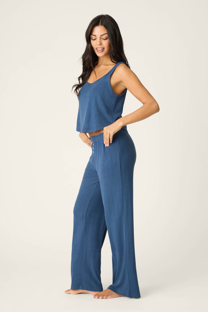 A woman stands barefoot against a plain background, wearing the P.J. Salvage Pointelle Essentials Deep Sea PJ Set with a blue cropped tank and matching wide-leg pants. She faces sideways, one hand on her hip, smiling down.