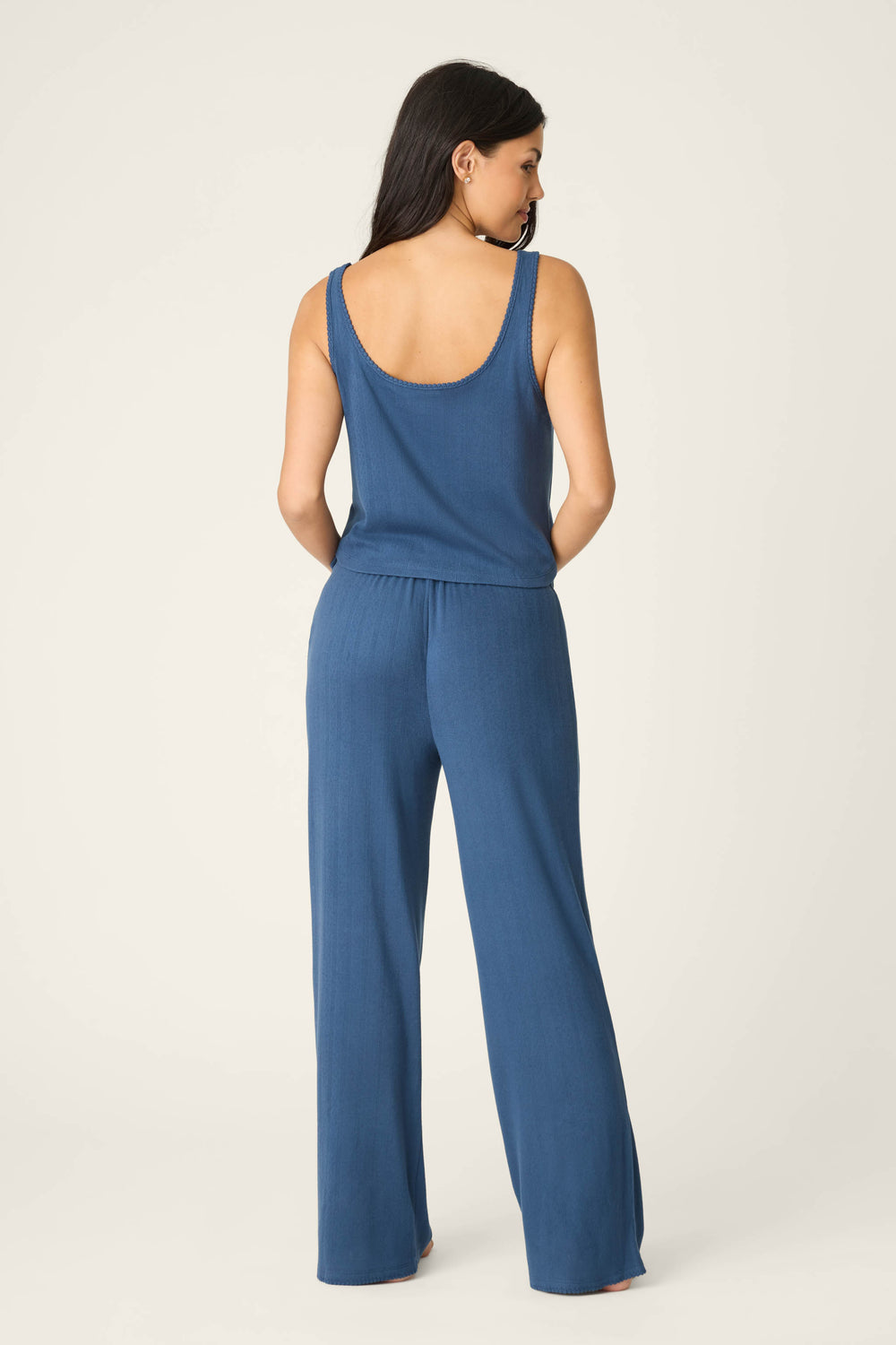 A person faces away from the camera in a P.J. Salvage Pointelle Essentials Deep Sea PJ Set, featuring a blue sleeveless scoop-neck top and matching wide-leg pants, against a plain light background.