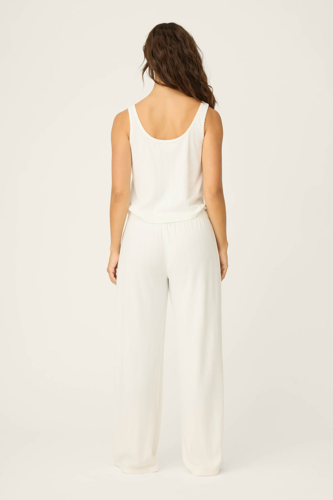 A woman with wavy brown hair, seen from behind, wears the sleeveless white Pointelle Essentials Ivory PJ Set by P.J. Salvage against a plain, light background.