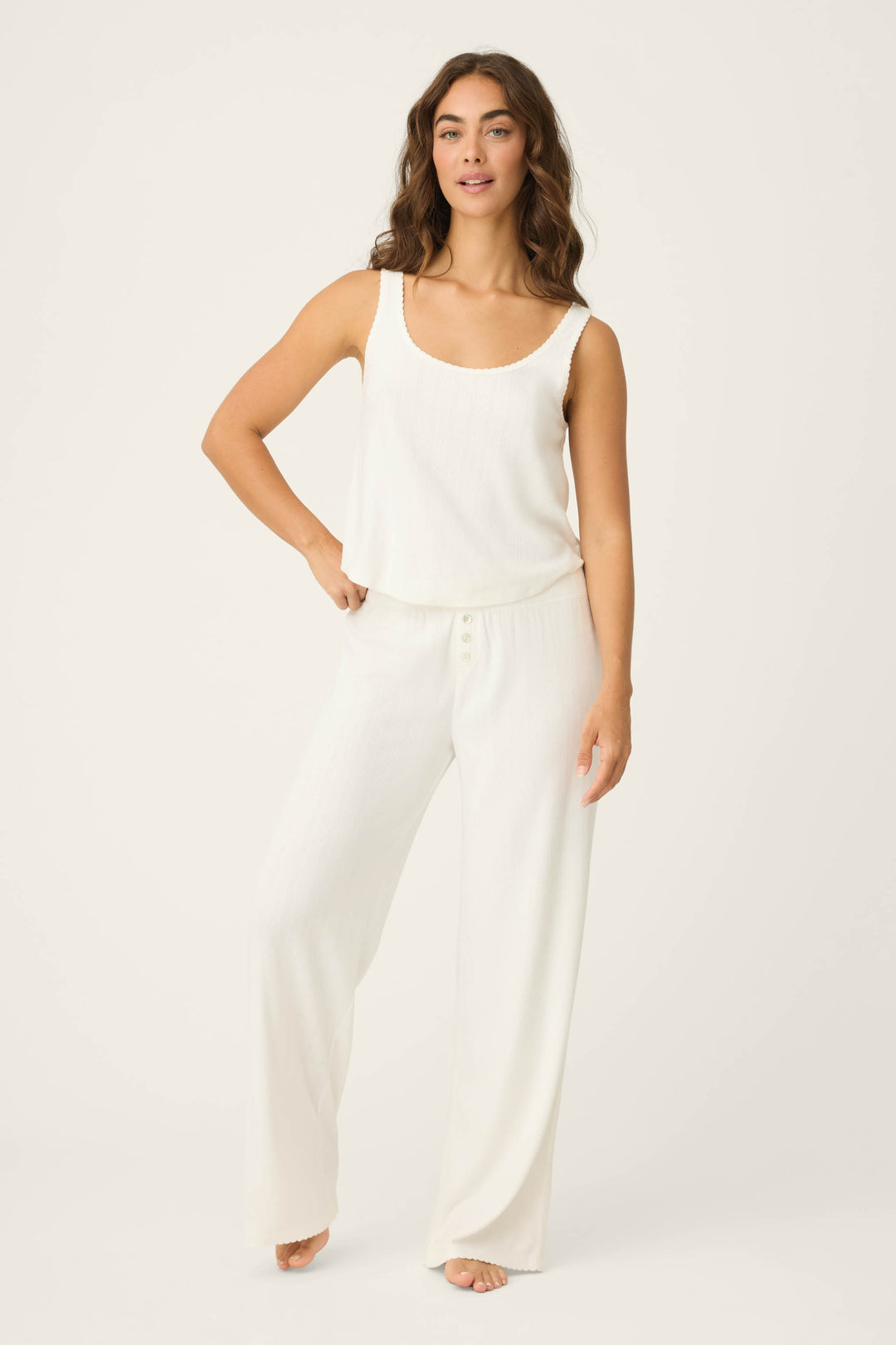 A person with wavy brown hair wears the Pointelle Essentials Ivory Tank Top by P.J. Salvage and loose white pants, standing barefoot on a light background with one hand on their hip.