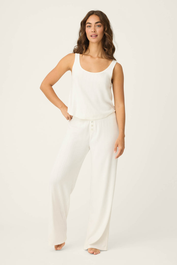 A person with wavy brown hair wears the Pointelle Essentials Ivory Tank Top by P.J. Salvage and loose white pants, standing barefoot on a light background with one hand on their hip.