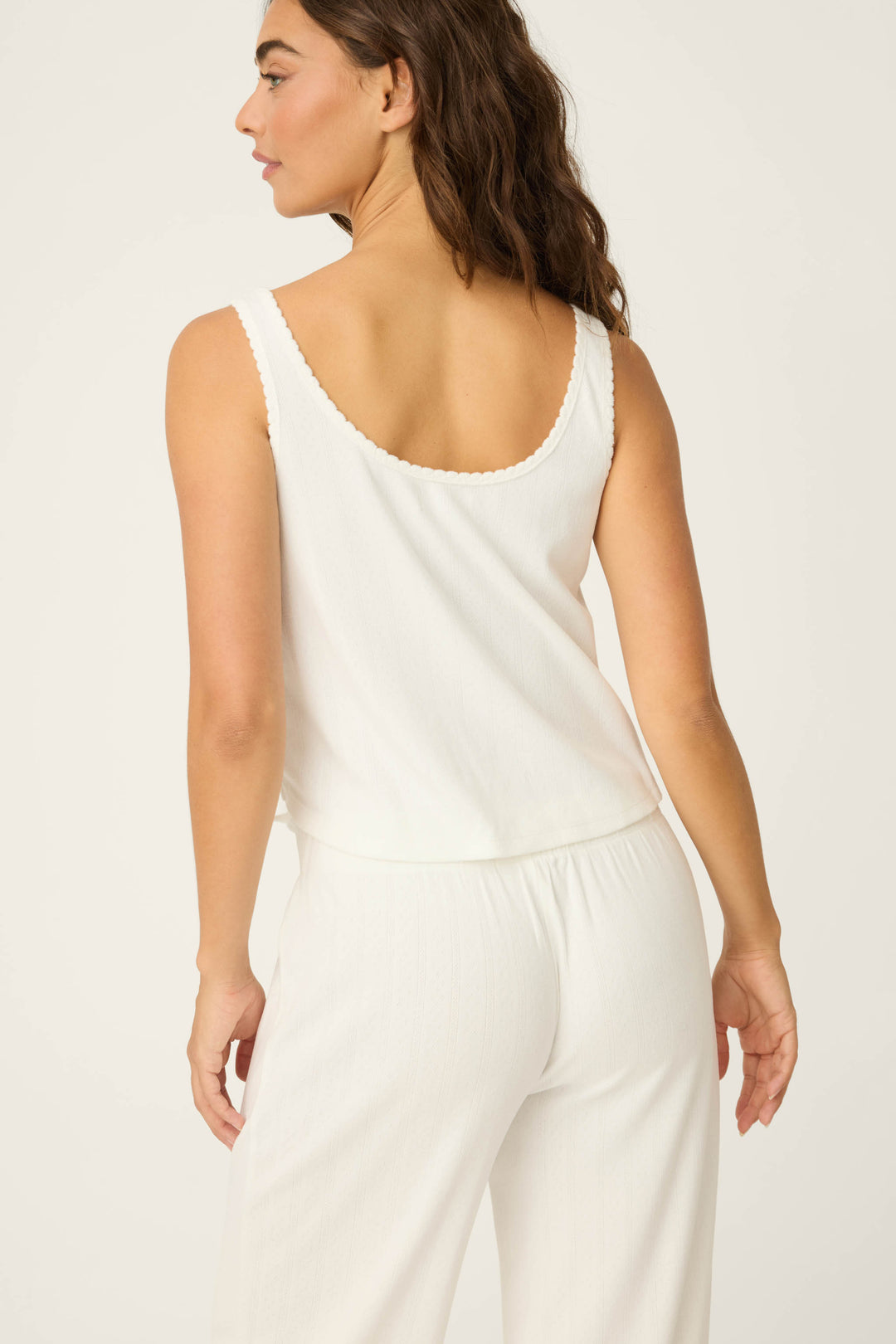 A woman with long brown hair is shown from the back, wearing the P.J. Salvage Pointelle Essentials Ivory Tank Top, paired with loose white pants, against a plain off-white background.