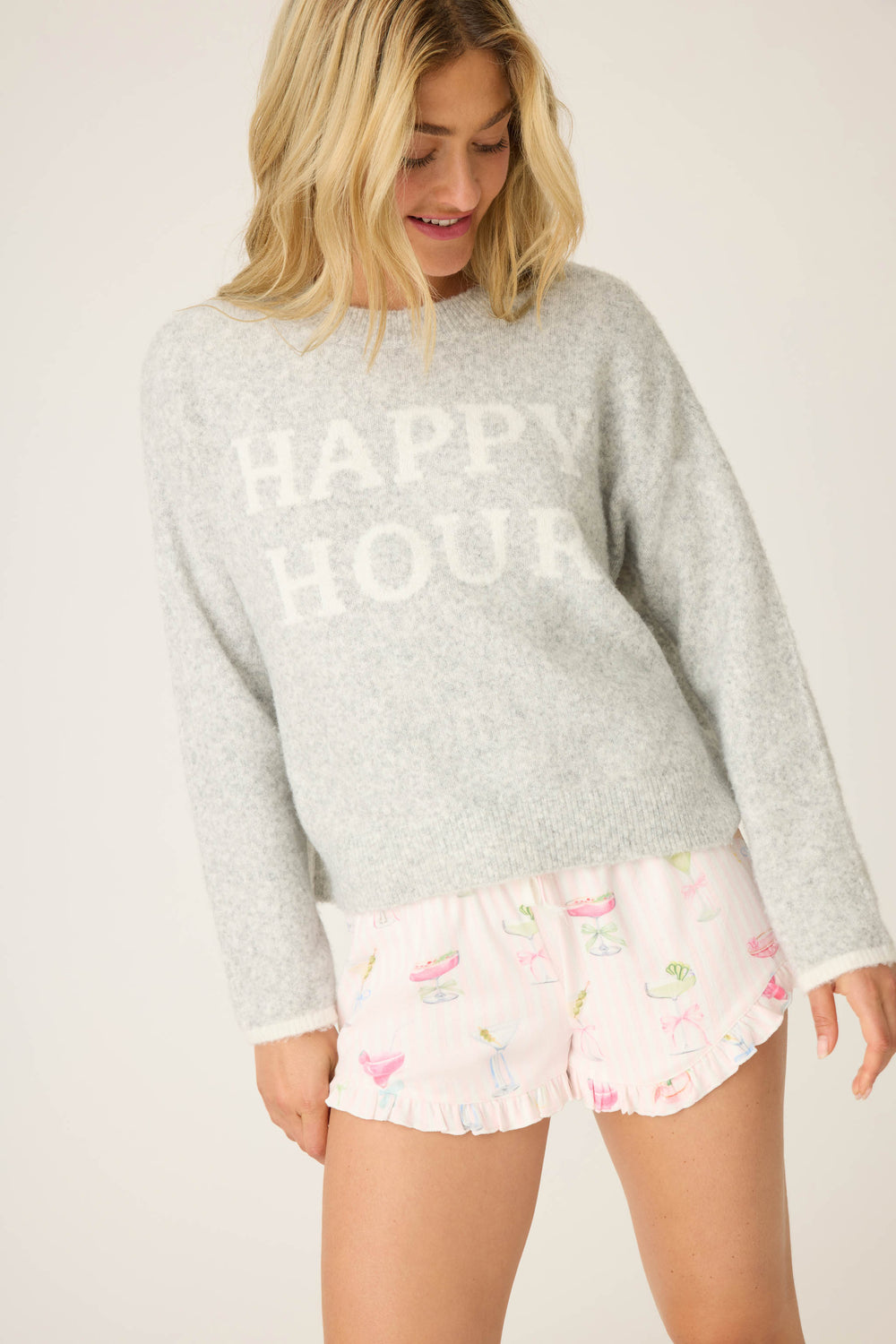 A blonde woman smiles while looking down, wearing the P.J. Salvage Pretty Punch Happy Hour Snuggle Sweater in light gray and pink pajama shorts with cocktails, donuts, and ruffled edges.
