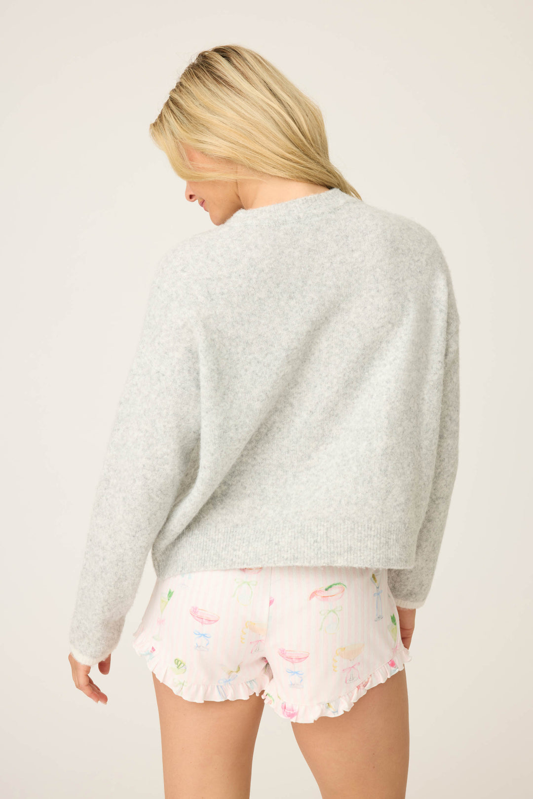 A person with long blonde hair faces away from the camera, wearing the P.J. Salvage Pretty Punch Happy Hour Snuggle Sweater in light gray and white ruffled pastel-print shorts, against a plain off-white background.