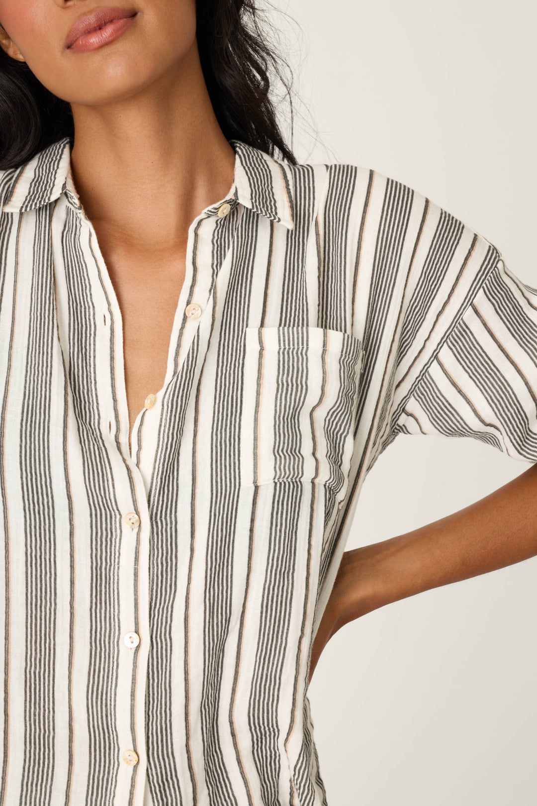 Woman models the P.J. Salvage Sea You in St. Tropez Shoreline Stripe Long Sleeve Top, featuring a relaxed fit, vertical black and white stripes, button front, and a front pocket. Her face is partly visible with her left hand on her hip.