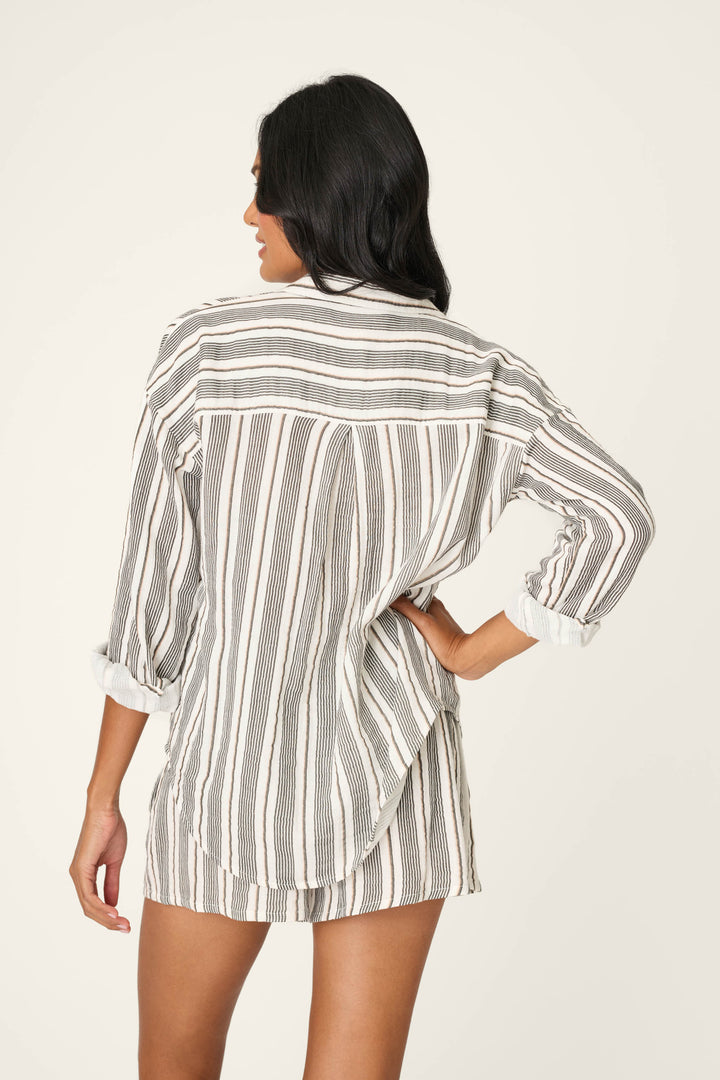 A woman stands with her back to the camera, wearing the P.J. Salvage Sea You in St. Tropez Shoreline Stripe Long Sleeve Top. One hand rests on her hip, her dark hair is loose, and the background is neutral.