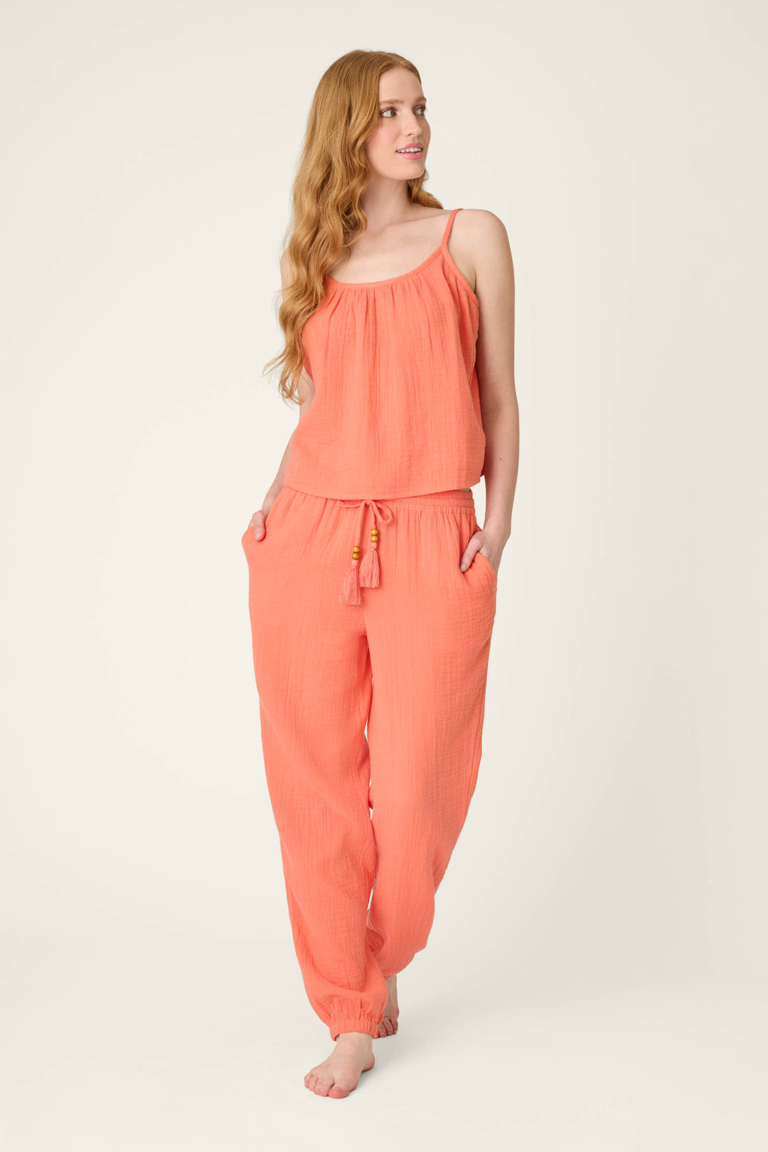 A person with long wavy hair stands barefoot, wearing the P.J. Salvage Sea You in Capri Hot Coral Cami with matching loose drawstring pants, hands in pockets, gazing slightly to the side.
