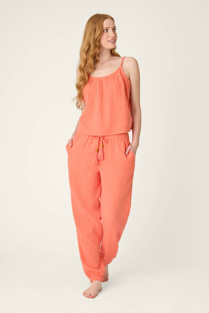 A person with long wavy hair stands barefoot, wearing the P.J. Salvage Sea You in Capri Hot Coral Cami with matching loose drawstring pants, hands in pockets, gazing slightly to the side.