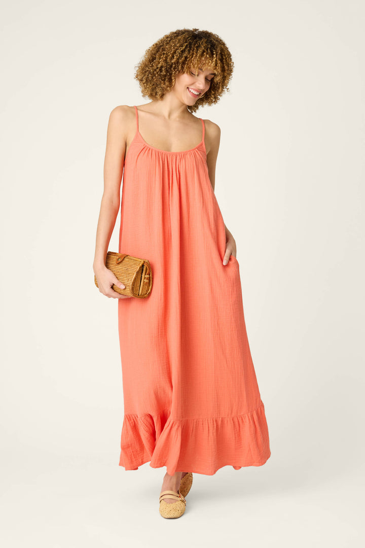A person with curly hair wears a sleeveless hot coral maxi dress with thin straps and ruffled hem, paired with tan accessories.