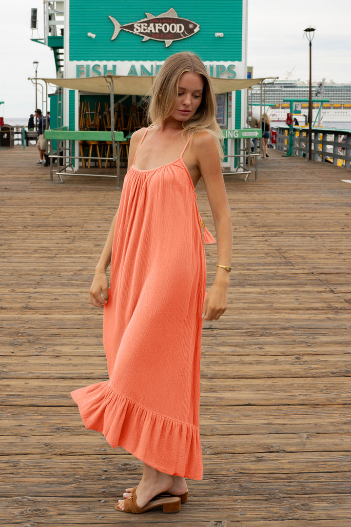 A woman in a bright coral dress and brown sandals stands on a wooden pier near a seafood restaurant, looking down.