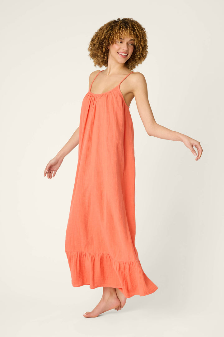 A person with curly hair stands barefoot against a plain light background, wearing a loose, sleeveless hot coral maxi dress with thin straps and a ruffled hem.