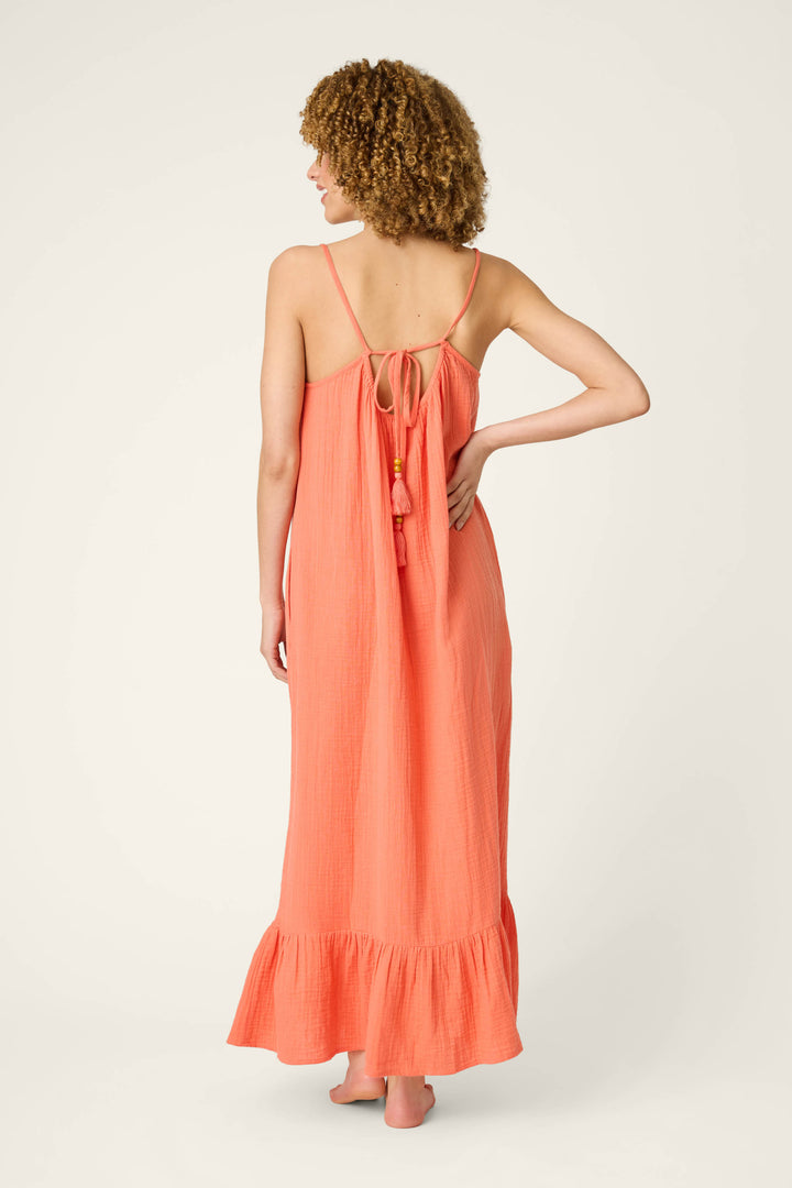 A person with curly hair stands barefoot, facing away, in a sleeveless hot coral maxi dress with thin straps and ruffled hem.