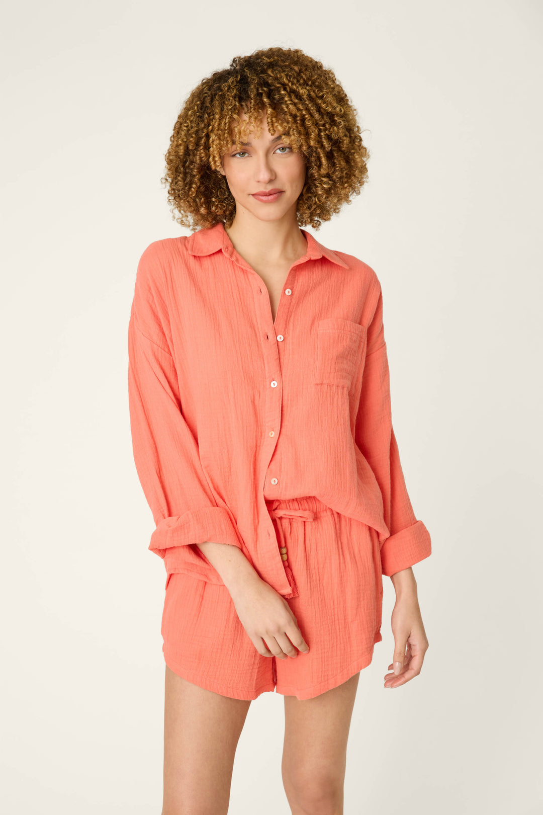 A person stands against a plain background wearing the P.J. Salvage Sea You in Capri Hot Coral Nightshirt with a front pocket. Their curly hair is styled naturally and their expression is neutral.