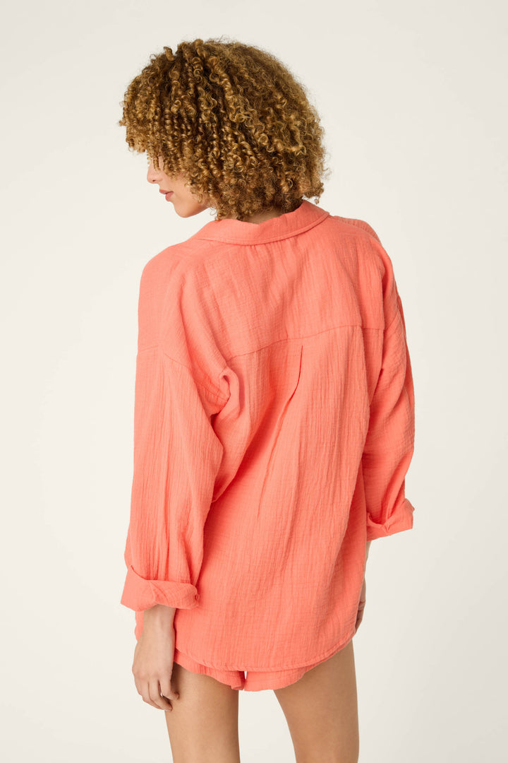 A person with curly hair faces away from the camera, wearing a loose-fitting hot coral nightshirt against an off-white background.