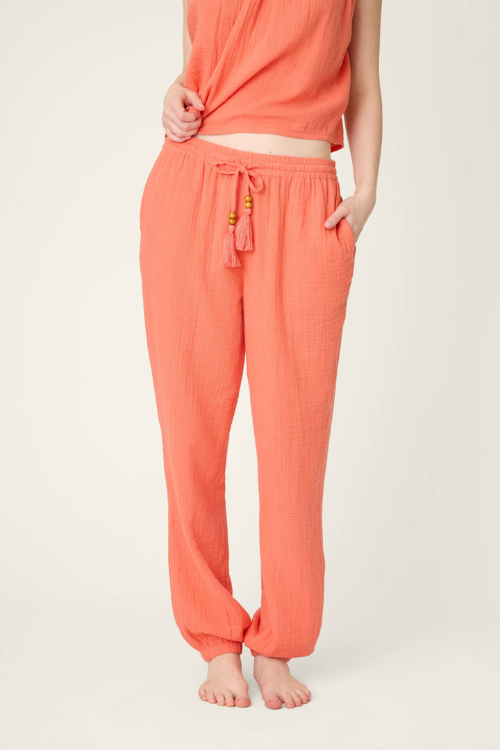 A person stands barefoot wearing hot coral pants with tassels and a drawstring, plus a matching sleeveless top, hands in pockets.
