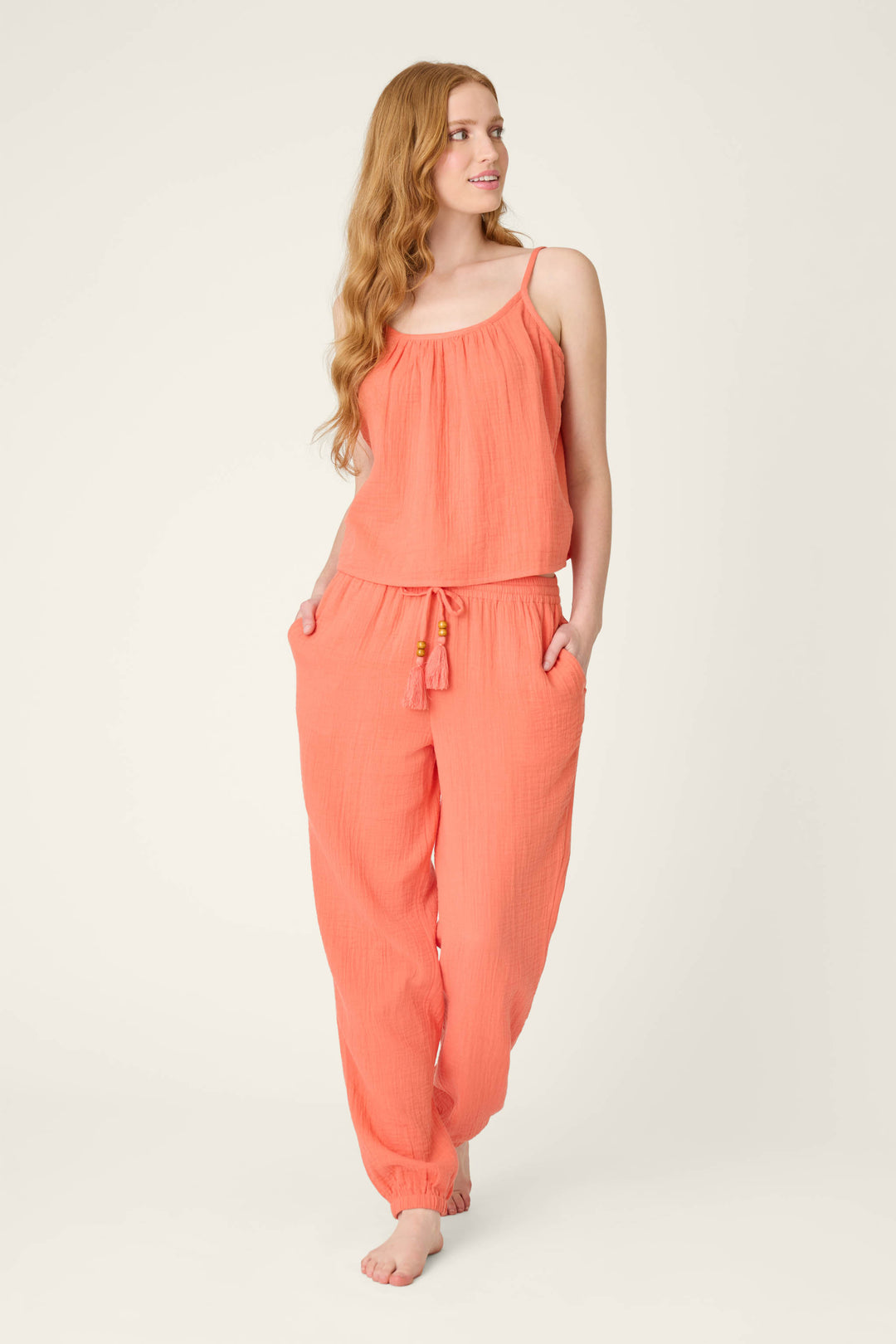 A woman with long, wavy hair stands barefoot against a plain background, wearing hot coral pants with her hands in the pockets, looking left.