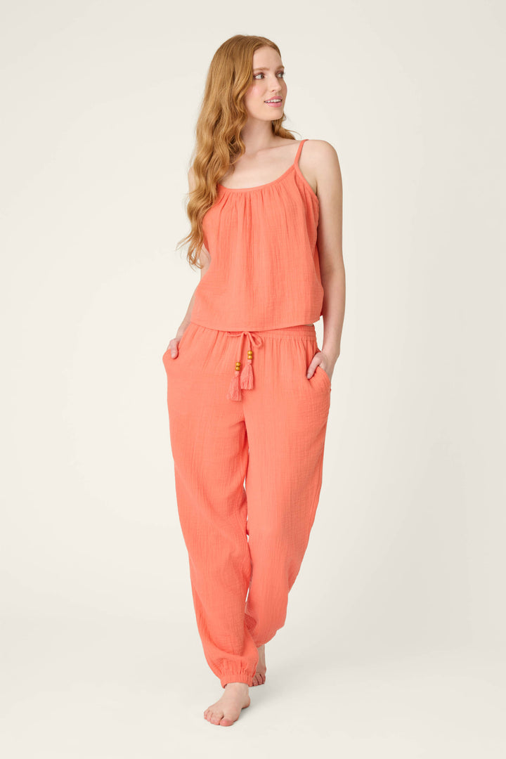A woman with long, wavy hair stands barefoot against a plain background, wearing hot coral pants with her hands in the pockets, looking left.