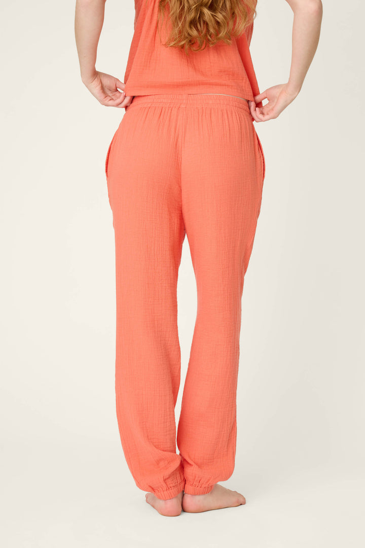 A person stands barefoot facing away, wearing hot coral pants and a matching top, lifting the top slightly at the waist against a plain light background.