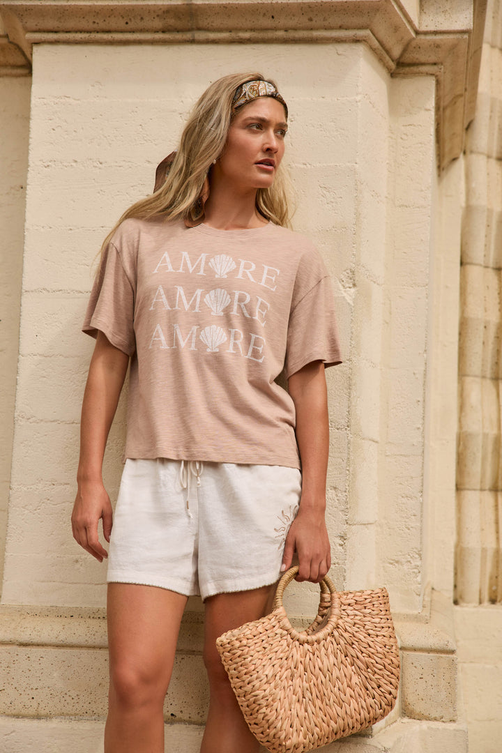 A woman stands against a stone wall wearing a beige T-shirt, white shorts, a headband, and holding a woven straw handbag.