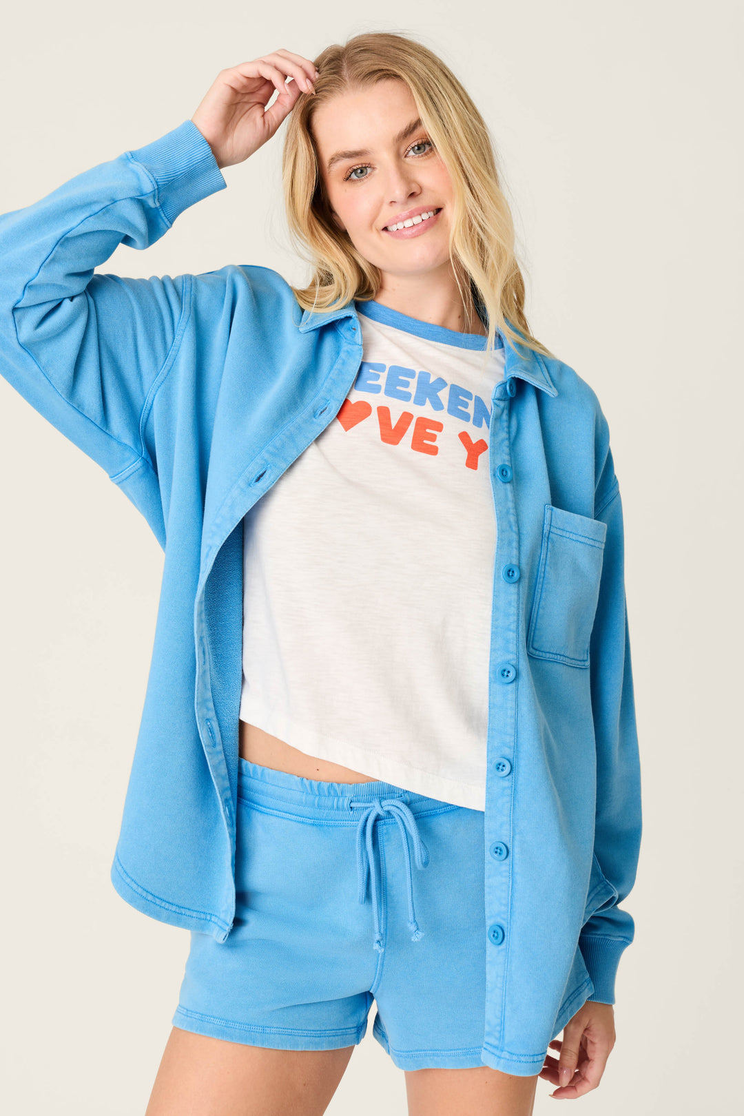 A woman smiles and touches her hair, wearing the P.J. Salvage Washed Away Marine Blue Shirt Jacket with matching shorts over a white crop top. The relaxed silhouette highlights her effortless style against a plain light background.