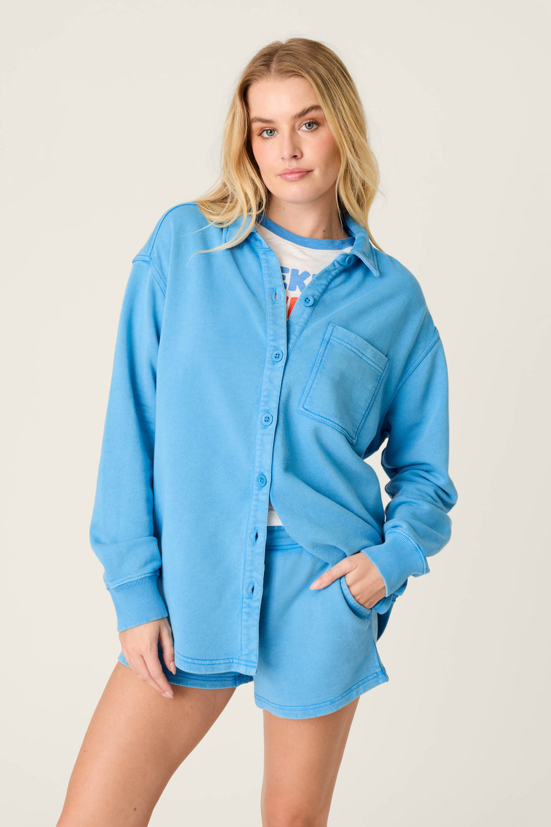 A person stands facing forward, wearing the P.J. Salvage Washed Away Marine Blue Shirt Jacket open over a printed tee, with one hand in the pocket of matching blue shorts, all set against a plain light background.