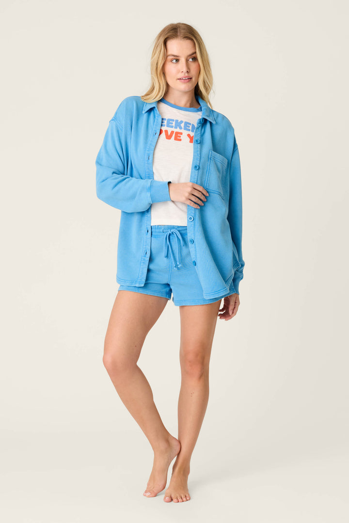 A woman stands barefoot against a plain background, wearing the P.J. Salvage Washed Away Marine Blue Shirt Jacket with matching shorts and a white T-shirt featuring WEEKEND LOVE in blue and orange, creating a relaxed silhouette.