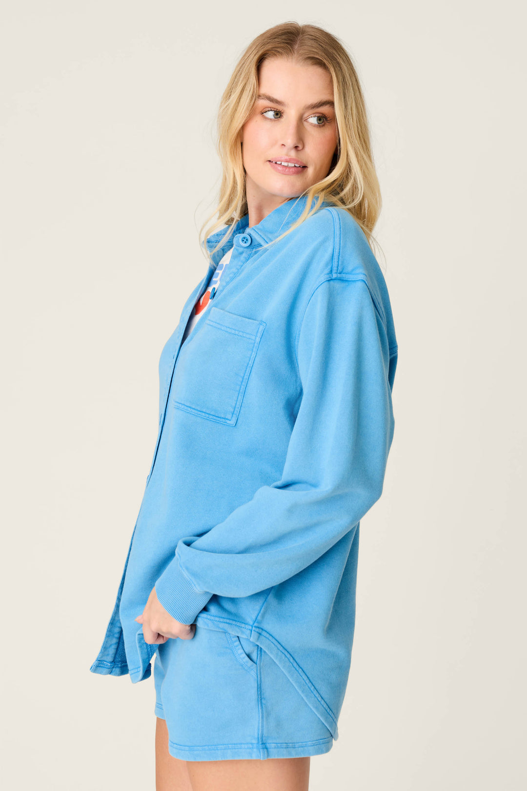 A person with long blonde hair wears the P.J. Salvage Washed Away Marine Blue Shirt Jacket with a chest pocket and a relaxed fit, paired with matching blue shorts, standing against a plain light background and looking to the side.