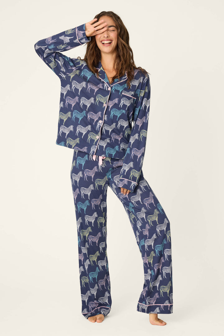 A woman stands barefoot, smiling and touching her forehead while wearing the P.J. Salvage Whimsy Technicolor Safari PJ Set: matching navy blue pajamas with a colorful zebra print, button-up top, and loose-fitting pants.