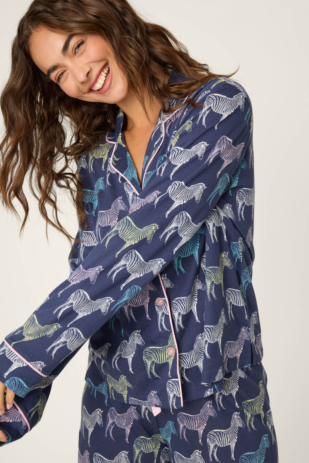 A woman with long wavy hair smiles while wearing the P.J. Salvage Whimsy Technicolor Safari PJ Set, featuring a colorful zebra print on dark blue fabric. She poses against a plain, light-colored background.