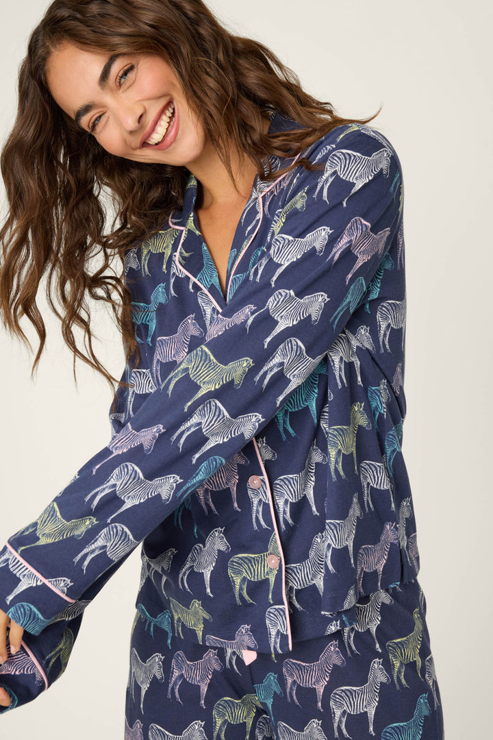 A woman with long wavy hair smiles while wearing the P.J. Salvage Whimsy Technicolor Safari PJ Set, featuring a colorful zebra print on dark blue fabric. She poses against a plain, light-colored background.