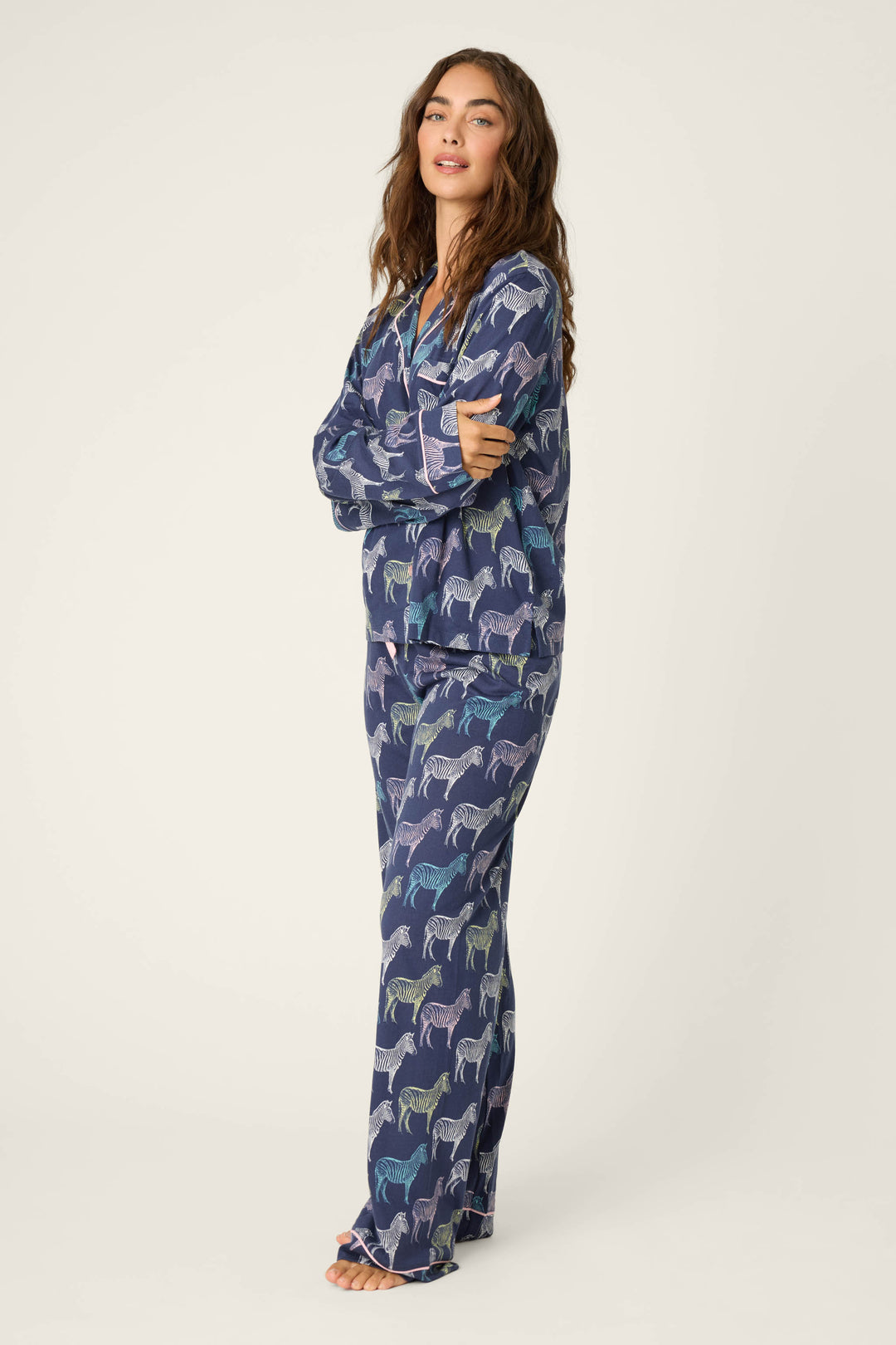 A person stands barefoot, arms crossed, wearing the P.J. Salvage Whimsy Technicolor Safari PJ Set—relaxed blue pajamas with a colorful zebra print—against a neutral background.
