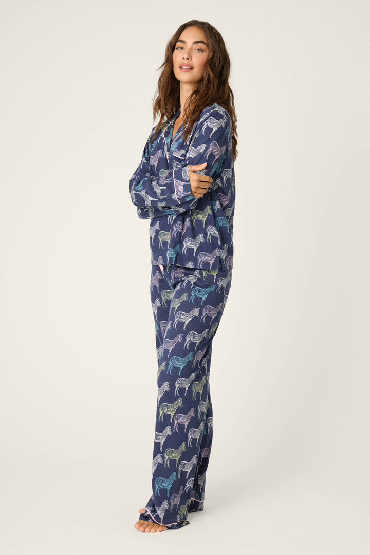 A person stands barefoot, arms crossed, wearing the P.J. Salvage Whimsy Technicolor Safari PJ Set—relaxed blue pajamas with a colorful zebra print—against a neutral background.