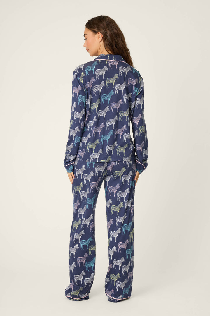 A person stands with their back to the camera, wearing the P.J. Salvage Whimsy Technicolor Safari PJ Set—a matching long-sleeve pajama set with a multicolored zebra print on dark blue—against a plain, light background.